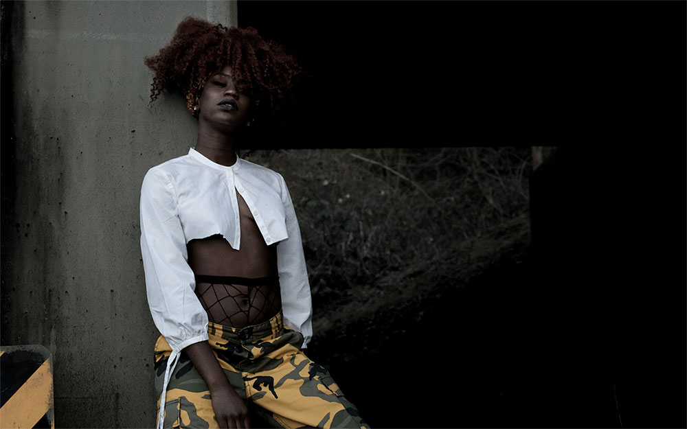 Black woman with curly, maroon hair 
            standing with her back to a stone wall. Her legs are slightly bent and she is facing the camera.
            Woman is wearing yellow and black camouflage pants and a white cut-off button up shirt.