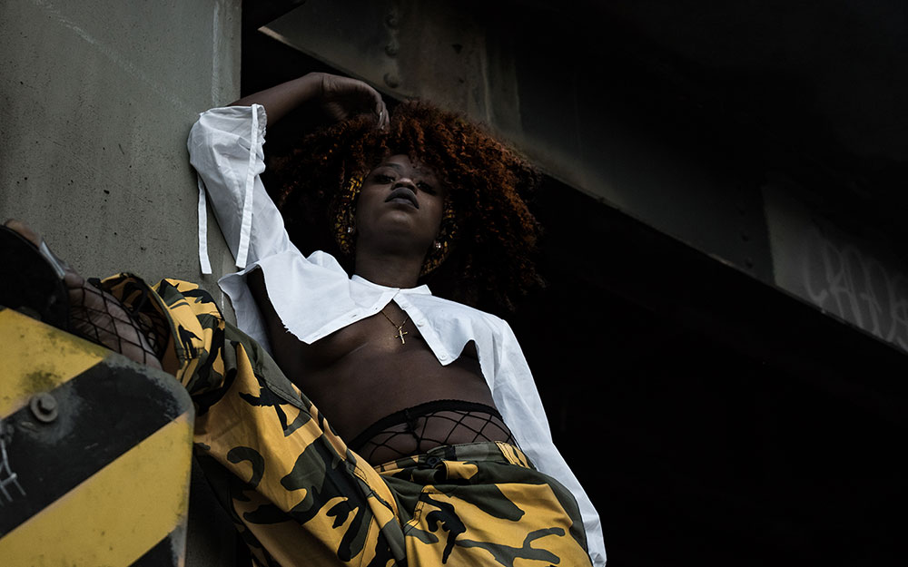 Black woman with curly, maroon hair 
            standing above caution sign under a dark underpass. Woman is wearing yellow and black camouflage pants.