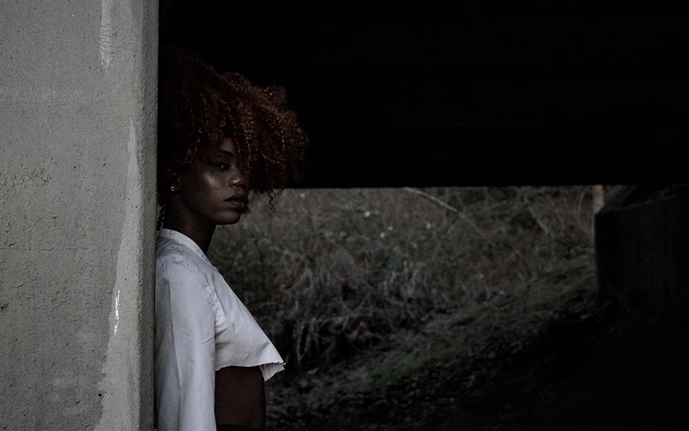 Black woman with curly, maroon hair standing 
            with her back to a stone wall. Her body is faced to the right, while her 
            head is turned towards the camera.