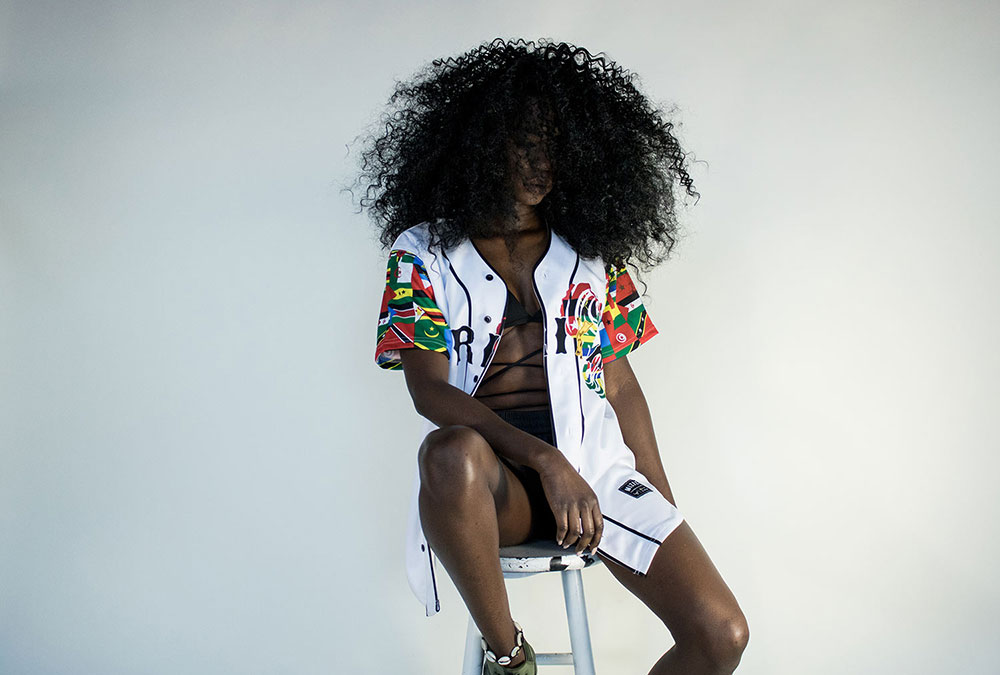 Black woman with curly, black hair sitting on a stool
            wearing a multicolored baseball jersey.The background is white.