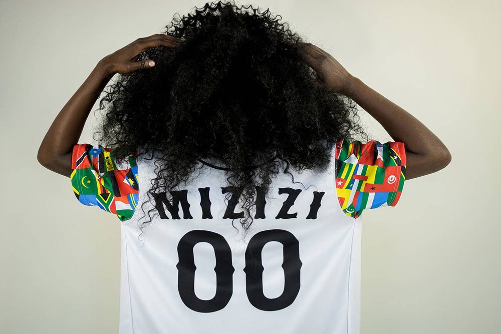 Black woman with curly, black hair wearing a 
            multicolored baseball jersey standing with their back to the camera and hands in their hair. The background is white.