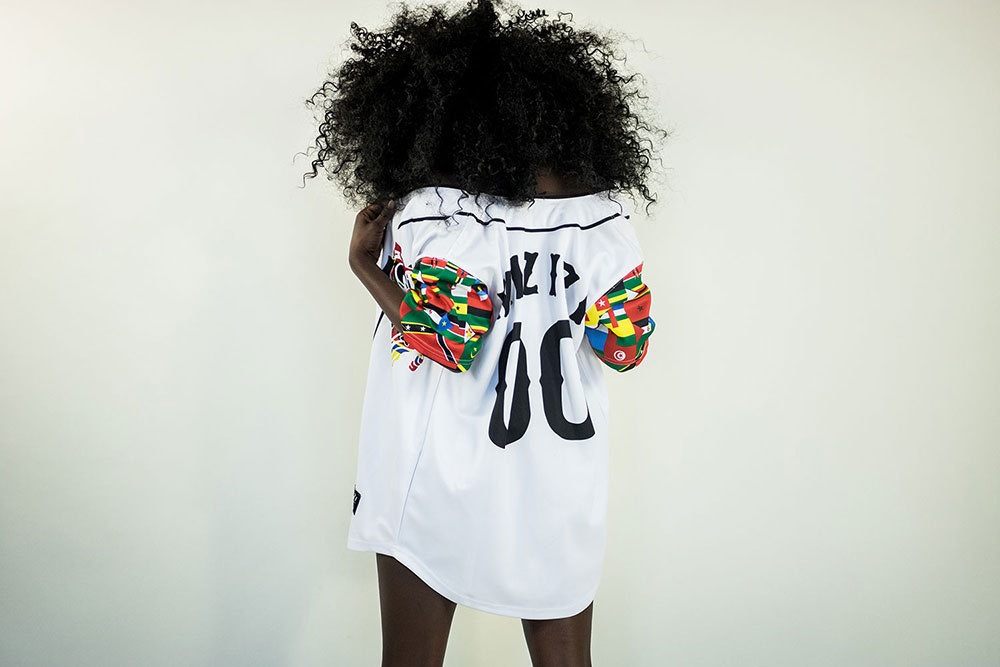Black woman with curly, black hair wearing a multicolored baseball 
            jersey standing with their back to the camera. The background is white.