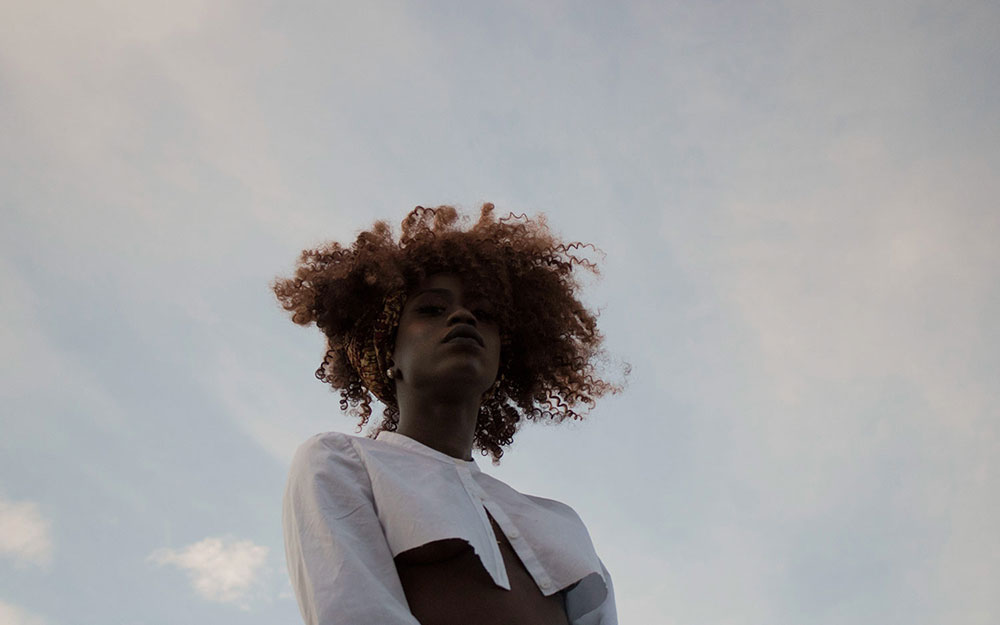 Portrait of maroon-haired black woman modeling white crop-top shirt in front 
                    of grey skies.
