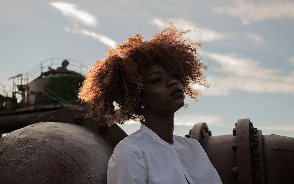 Black woman with curly, maroon hair looking out into the 
                distance. Behind the woman are maroon pipes and gray skies.
                The woman is wearing a white button-up shirt.