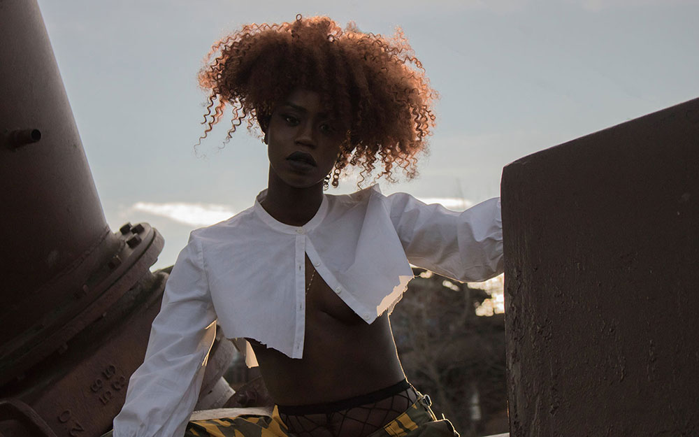 Black woman with curly, maroon hair crouching between 
                maroon pipes. The woman is staring directly into the camera. The woman is wearing a white button-up shirt 
                and yellow and black camouflage pants.