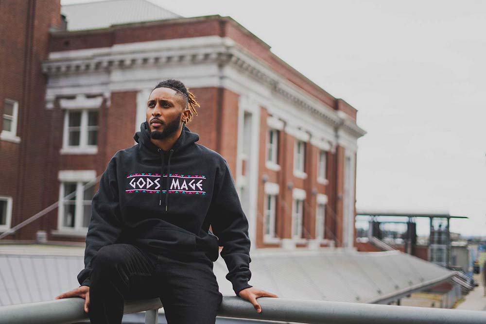 Man sitting on railing modeling black hoodie that reads 'Gods Image'. 
            Man's torso facing camera while he looks out into 
            the distance.