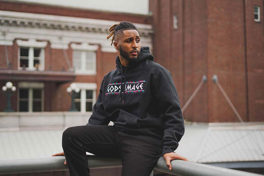 Man sitting on railing modeling black hoodie that reads 'Gods Image'.
            Man's torso is facing camera, while head is turned to the left.