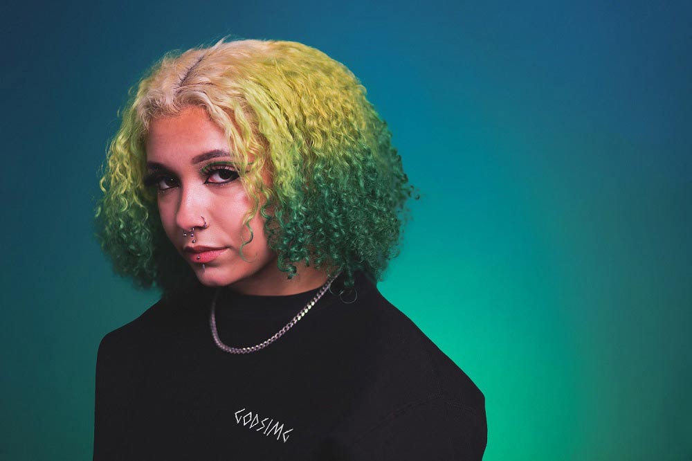 Person modeling black 'GODSIMG' crewneck. Person has curly, green and white hair and is gazing out towards 
                the distance.
