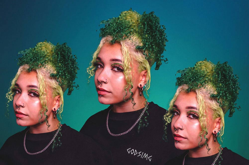 Person modeling black 'GODSIMG' crewneck. Special effect makes person appear three times side by side. 
                Person has curly, green and white hair and is looking directly at camera.