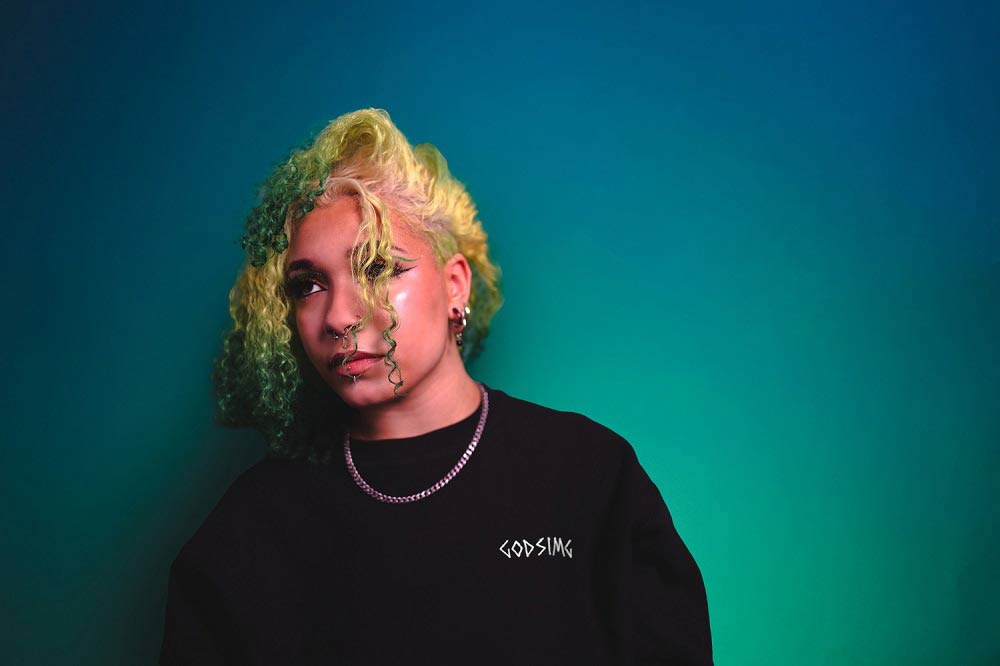Person wearing black crewneck which reads 'GODSIMG' across the left 
                    breast. Person has curly, green and white hair and is gazing out towards 
                    the distance.