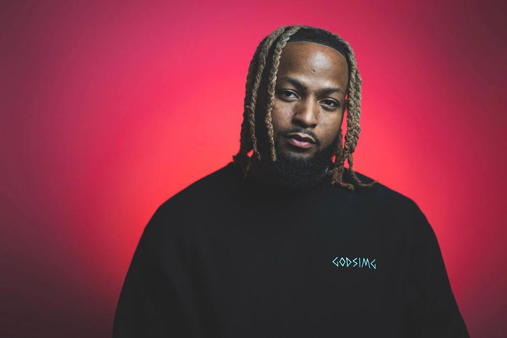 Man modeling black 'GODSIMG' crewneck in front of red and orange backdrop. 
                Man looking directly at camera.