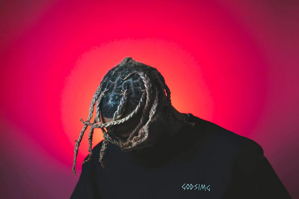 Man modeling black 
                'GODSIMG'crewneck in front of red and orange backdrop. Man's hair covering face.