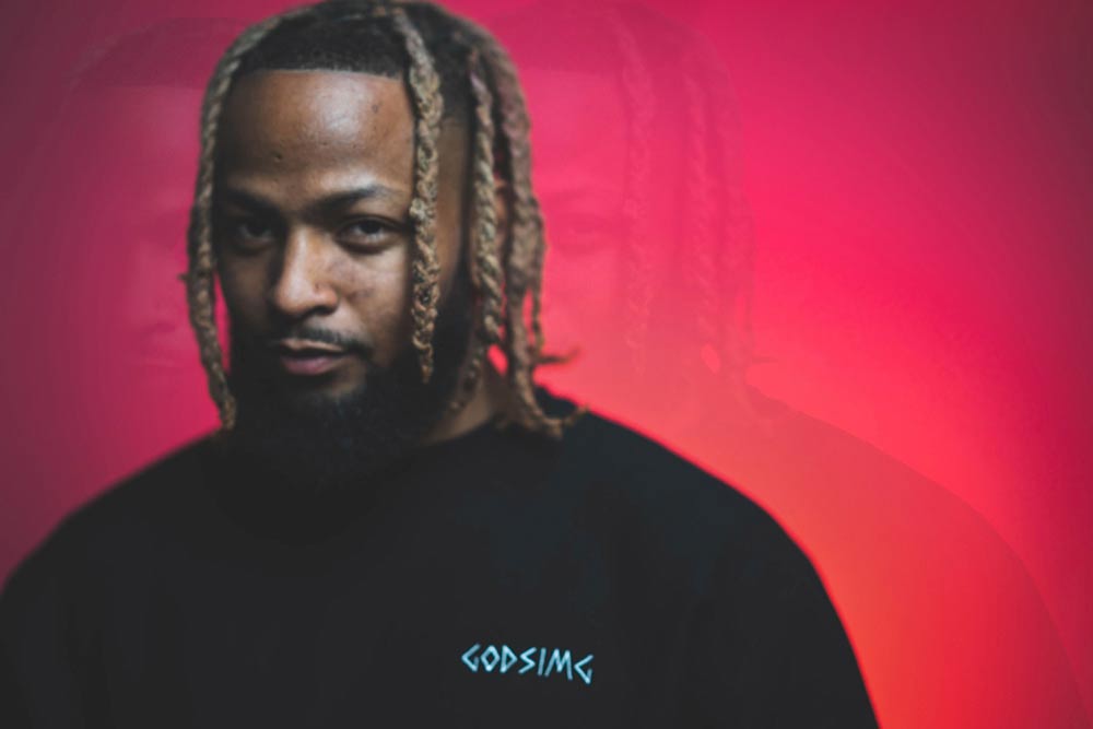 Man modeling black 
                'GODSIMG' crewneck in front of red and orange backdrop. Special effect 
                applied to make person appear two addional times. Man looking directly at camera.