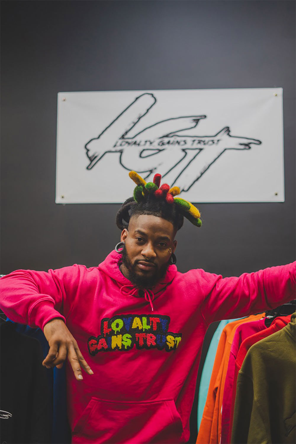 Man standing in between aisles of merchandise. His hair is styled
            in red, green and yellow dreadlocks and he is wearing a majenta hoodie that says Loyalty
            Gains Trust across the front. In the background is a white banner with the letters LGT displayed.