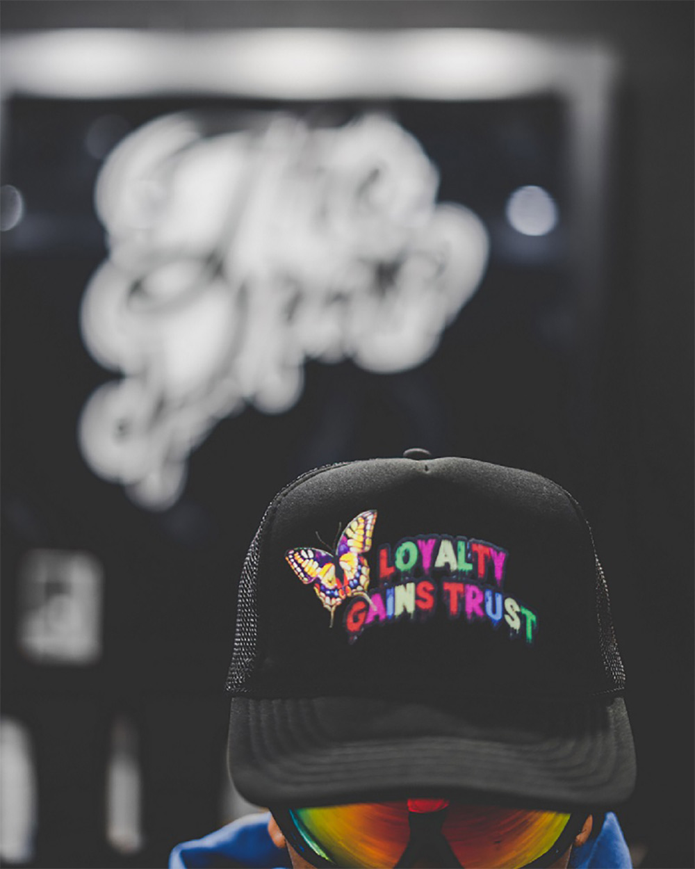 Man wearing black hat and sunglasses with red and yellow lenses. 
                    Hat has butterfly decoration next to text that reads 'Loyalty Gains Trust'.