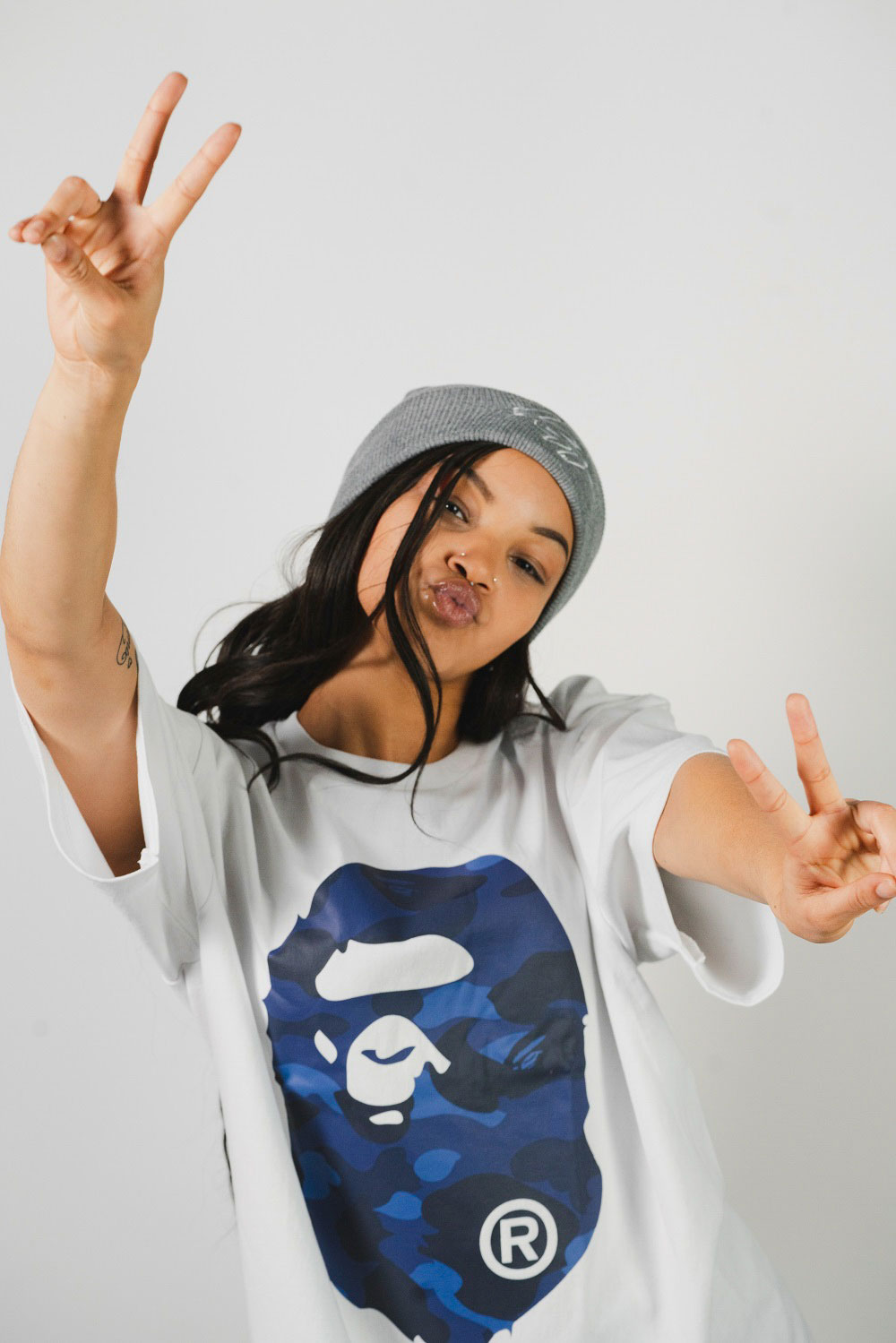 Woman wearing
            white t-shirt with blue Bape logo across the front of it, and a grey beanie.
            She is making piece signs with both of her hands.