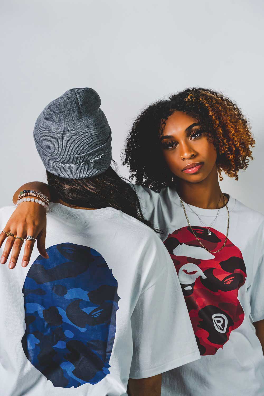Two women modeling Bape t-shirts. One is facing the camera and the other has their back turned to it. 
                        The t-shirt on the left displays a large, red Bape logo and t-shirt on the right 
                        displays large, blue Bape logo.
