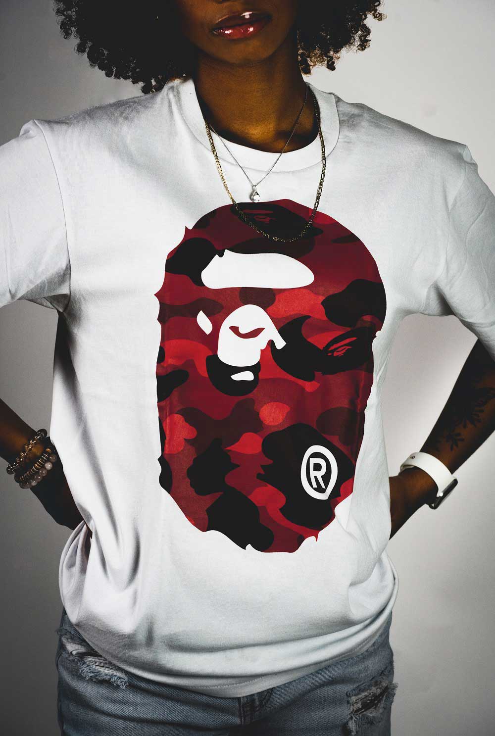 Curly-haired woman modeling
            white t-shirt with large, red Bape logo across front of it. She is standing with 
            hands on hips facing the camera. Photo spans from her lips to her hips.