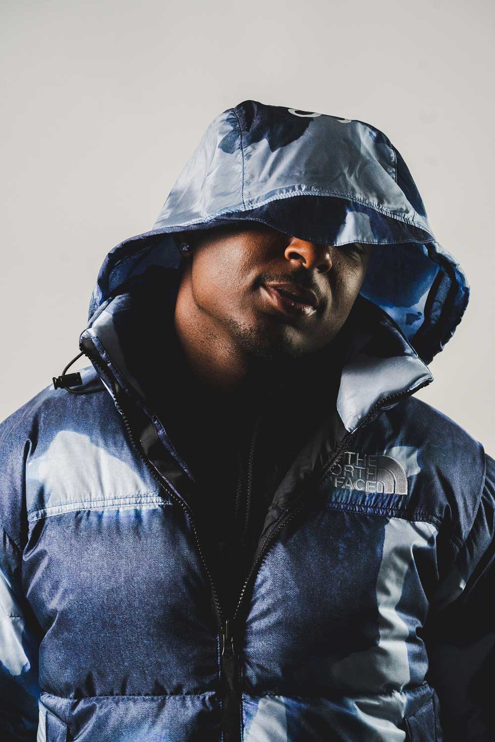 Man modeling
        blue camouflage North Face jacket. Man is facing camera and a smile appears across 
        man's face but rest of face is covered under hoodie.