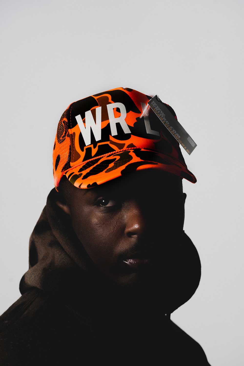 Man modeling
            orange and black camouflage hat. Front of hat reads 'WRLD'. Dark shadow cast 
            over part of hat's text and man's face.