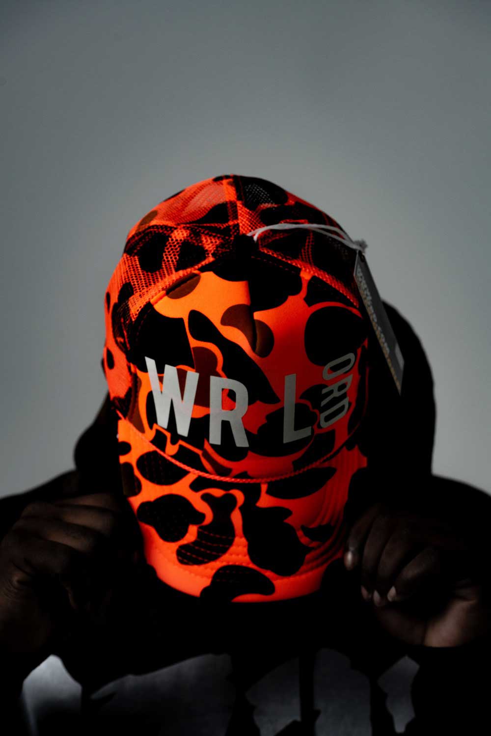 Man modeling
            orange and black camoflauge hat. Front of hat reads 'WRLD' and man
            is holding both sides of brim with head titled downwards. 