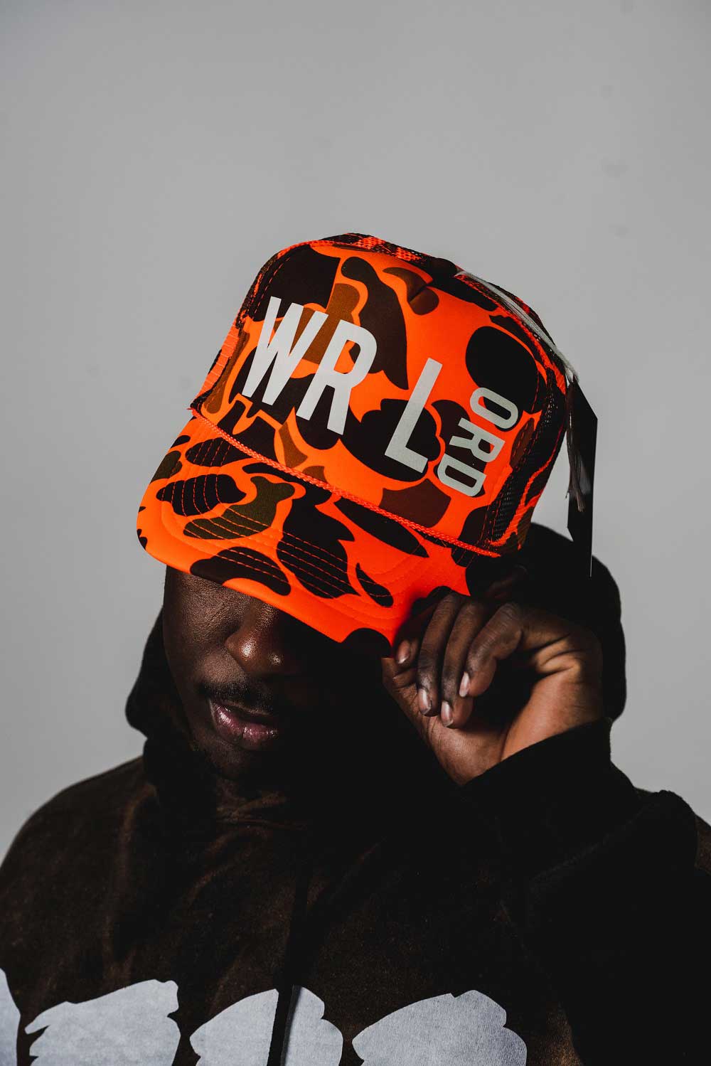 Man modeling
            orange and black camouflage hat. Front of hat reads 'WRLD'in white 
            text. Man holding brim of hat.